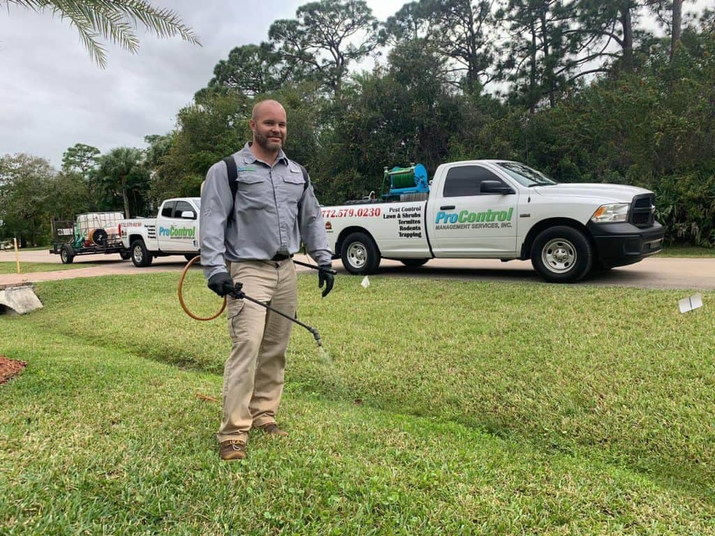 A person wearing work attire is standing on a grassy lawn, holding a spraying device in one hand. Two white work trucks with "ProControl" logos and attached utility trailers, clearly part of a pest control service, are parked on a street in the background, surrounded by trees and greenery.