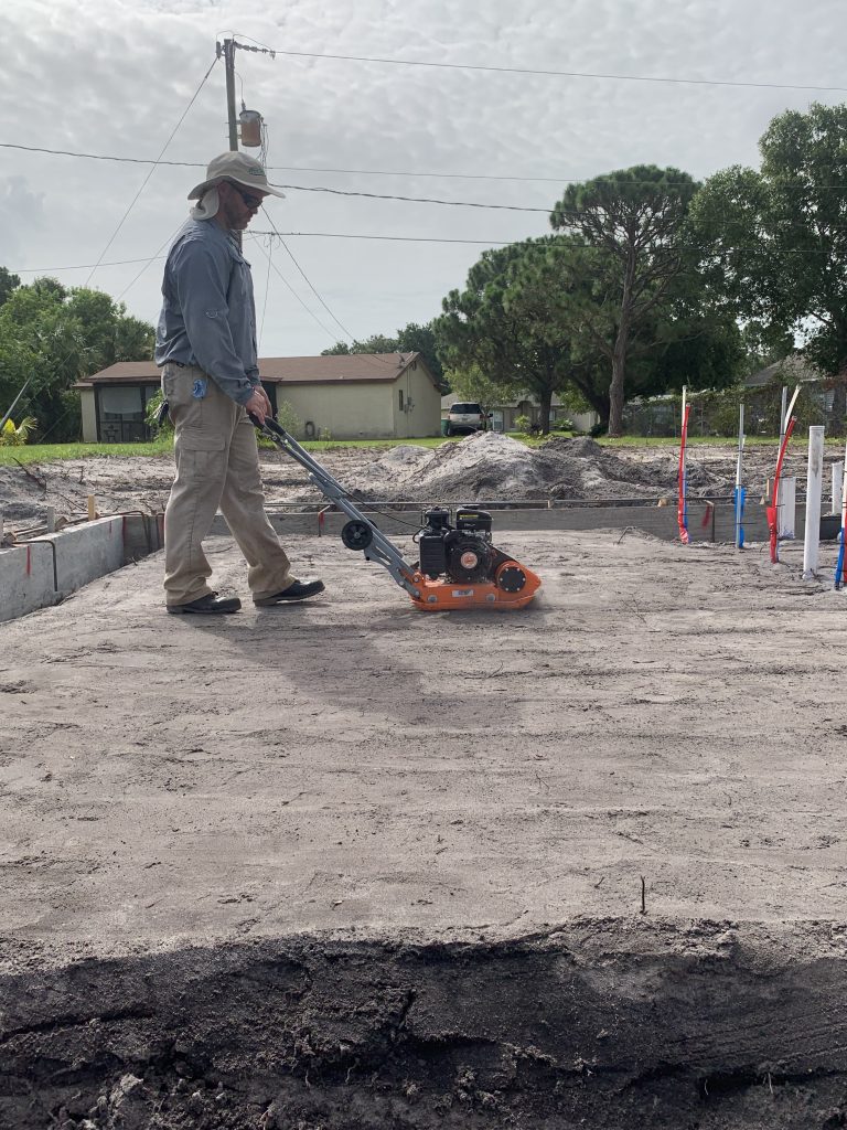 A person wearing a hat, sunglasses, and long sleeves operates an orange plate compactor on a sandy construction site with plumbing pipes and houses in the background—just one example of the dedication found in our About Us story.
