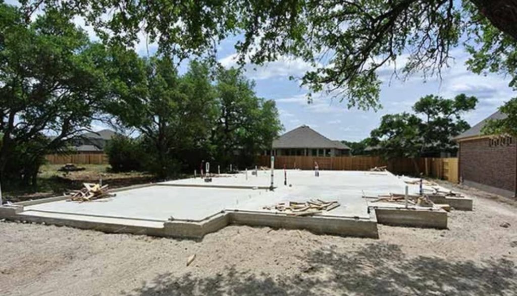 About Us: Laying a concrete foundation for a new house, surrounded by dirt, trees, and nearby homes. Plumbing pipes rise from the slab, with construction materials scattered across the site.