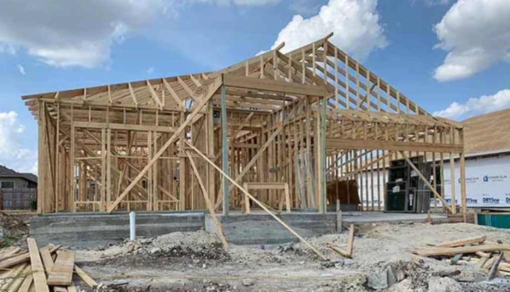 About Us: A wooden house frame under construction, with exposed beams and supports, stands on a concrete foundation. The sky is partly cloudy, and building materials are scattered around the construction site.