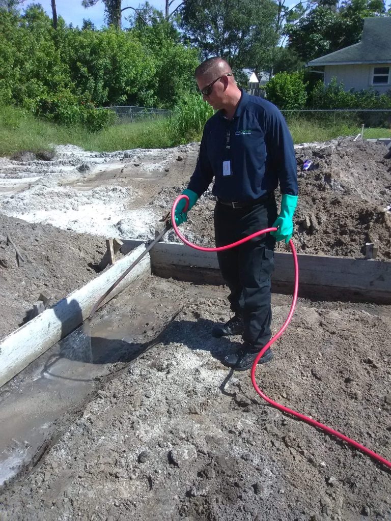 A person wearing gloves, sunglasses, and black clothing uses a red hose to spray water or chemicals on the ground at a construction site, with dirt, wooden beams, and greenery in the background—perfect for our About Us page.