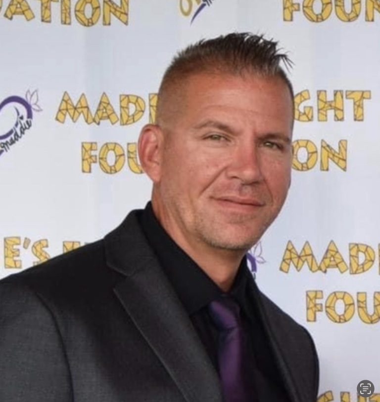 A man with short, spiked hair wearing a dark suit, black shirt, and dark tie stands in front of a step-and-repeat banner with yellow writing for "Madie's Fight Foundation" at an About Us event.
