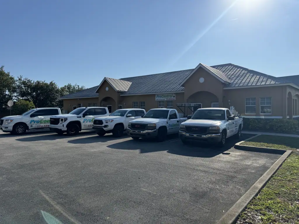 A row of white pickup trucks and SUVs with "ProCore" logos, offering pest control St Lucie, FL services, are parked in front of a single-story tan commercial building under a clear blue sky.