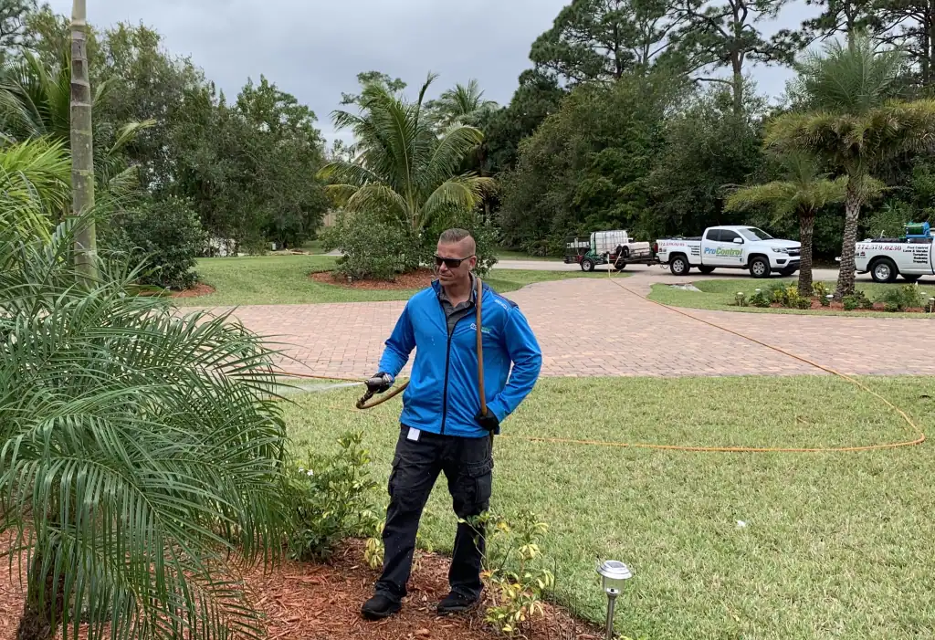 A man in a blue jacket and sunglasses sprays a palm plant for pest control St Lucie, FL. He holds a hose in a landscaped yard, with lawn, trees, and two white pickup trucks visible in the background.