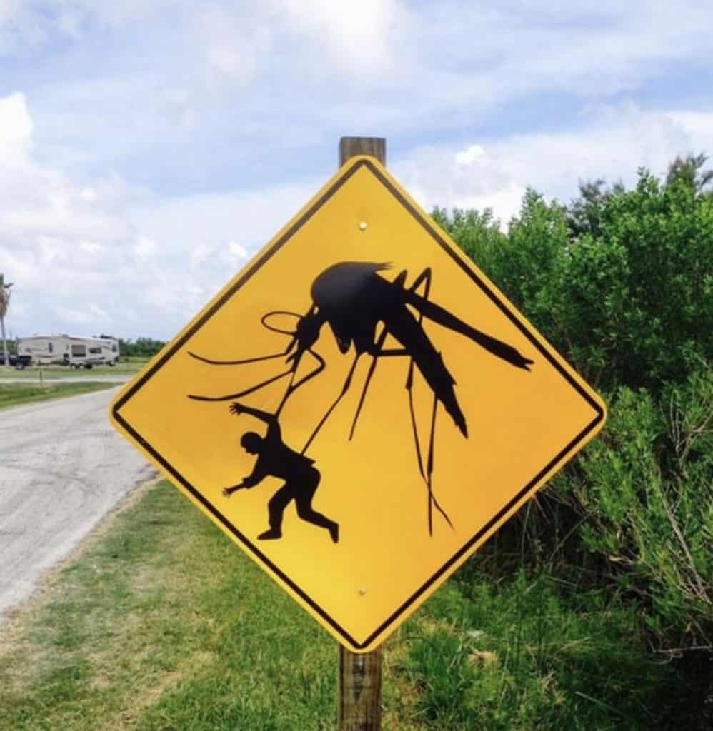A yellow warning sign in FL shows a giant mosquito grabbing a person, humorously suggesting large or aggressive mosquitoes in the area—perfect for reminding locals to consider pest control St Lucie. Trees and a road are visible in the background.