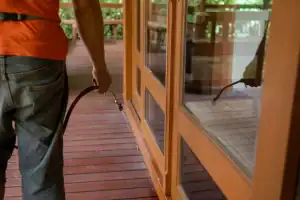 A person holding a spray nozzle is standing on a wooden deck near glass doors, possibly preparing for pest control St Lucie services. Only the person's arm and part of their body are visible in this FL scene.