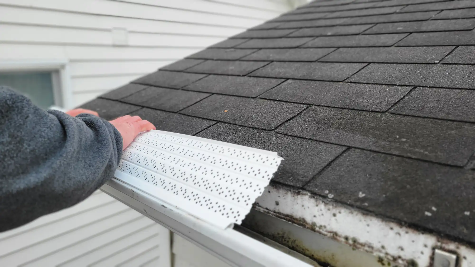 A person installs a white, perforated gutter guard on the edge of a shingled roof, next to a white house exterior.