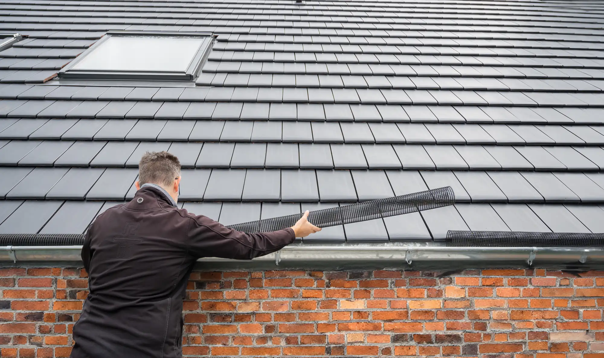 A person in a black jacket installs a gutter guard on a roof with dark gray tiles and a brick wall, standing below a skylight window.