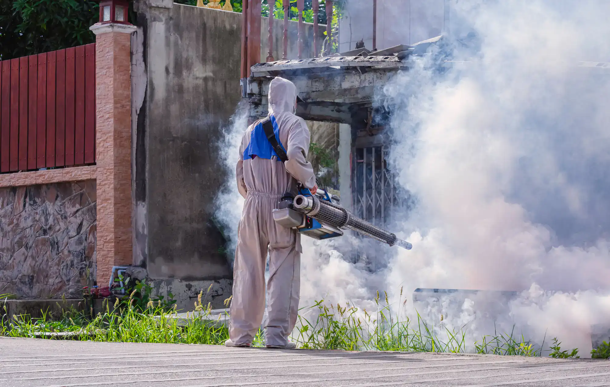 A person in protective clothing uses a fogging machine to spray insecticide outside a building, creating thick clouds of white smoke around them.