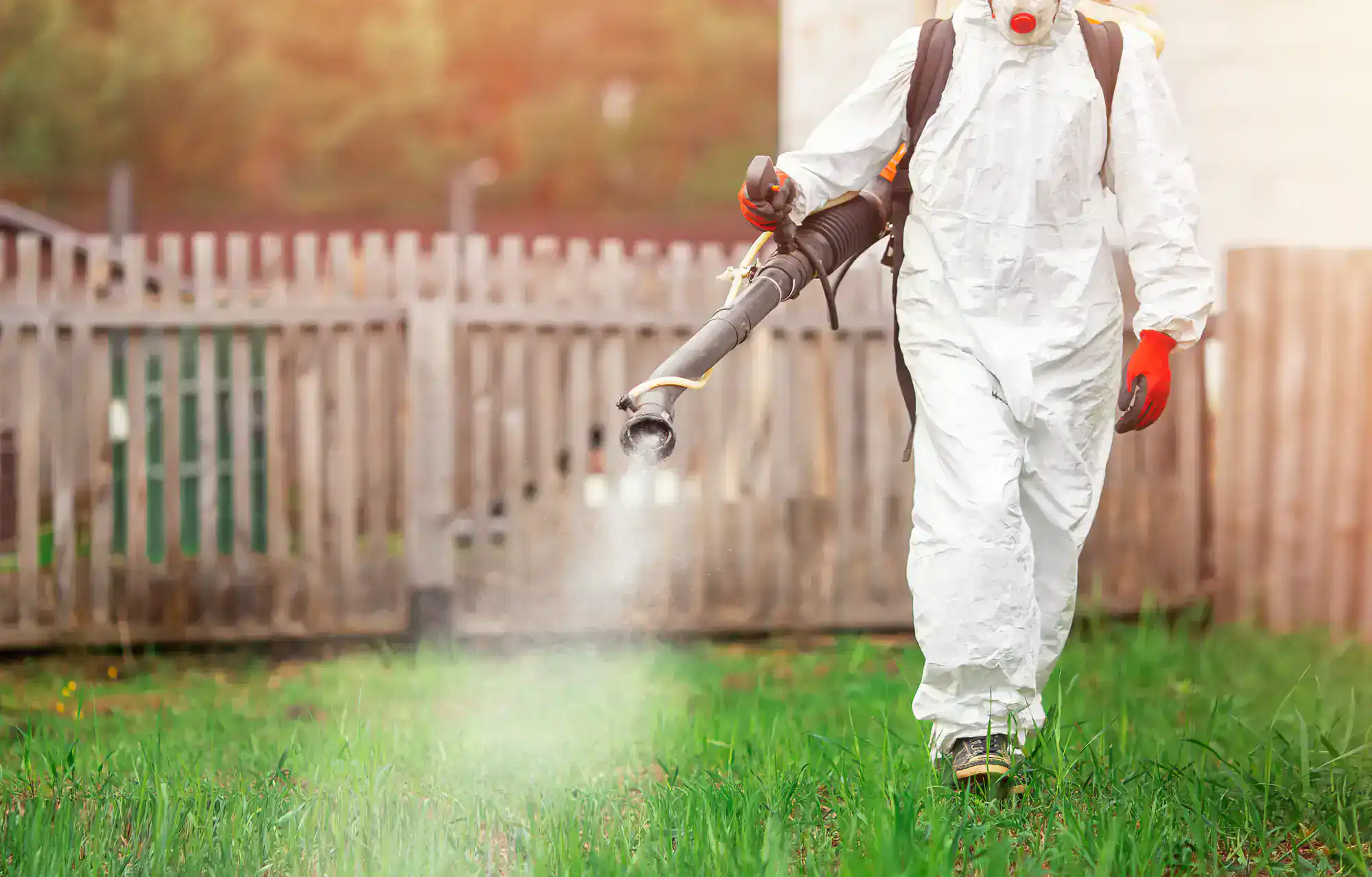 A person in a protective white suit, gloves, and mask sprays chemicals on grass with a backpack sprayer for pest control St Lucie, FL, in a grassy area near a wooden fence.