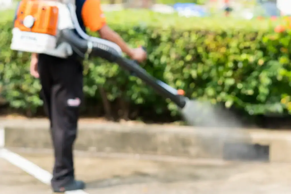 A person wearing an orange shirt and black pants uses a backpack leaf blower to clean a sidewalk near green bushes. The background is blurred.