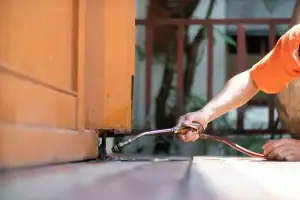 A person in an orange shirt uses a pest control sprayer to apply chemicals at the base of a wooden door, aiming to prevent or treat termite infestation.