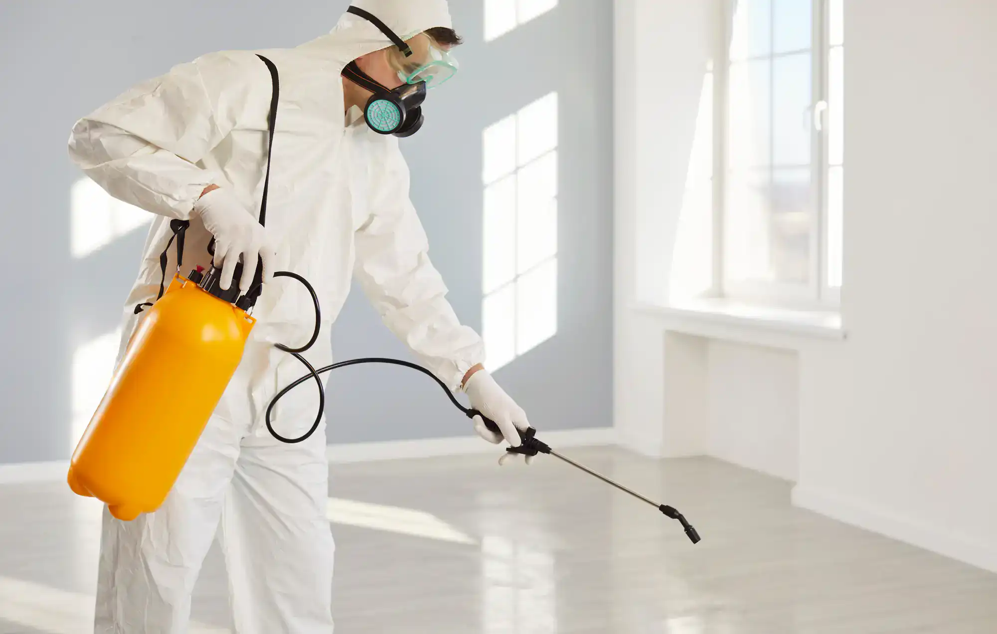A person wearing protective gear and a respirator mask uses a sprayer to disinfect or treat surfaces in a bright, empty room with large windows.
