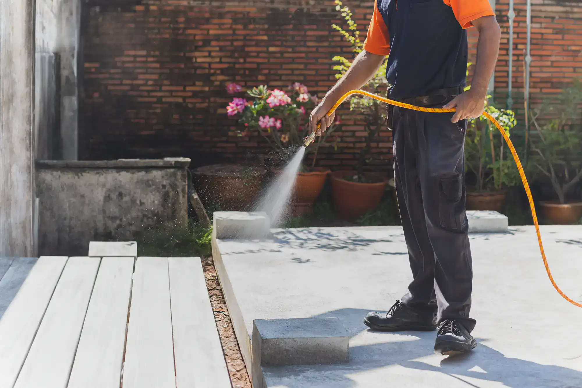 A person wearing dark pants and an orange shirt uses a garden hose to spray water on a paved outdoor area, with potted plants and a brick wall in the background.