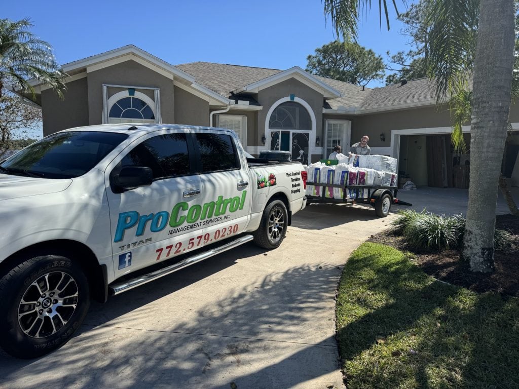 A ProControl Management Services pickup truck is parked in the driveway of a suburban house. A trailer loaded with Blown-In Insulation for Pests holds large bags, as a worker unloads items by the front entrance. Palm trees line the driveway.