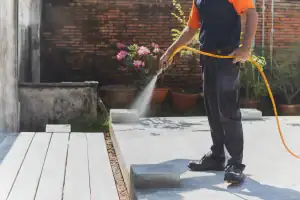 A person wearing dark pants and a short-sleeved shirt uses a yellow hose to spray liquid onto a concrete surface outside, possibly for Pest Control in St. Lucie County, with potted plants and a brick wall in the background.