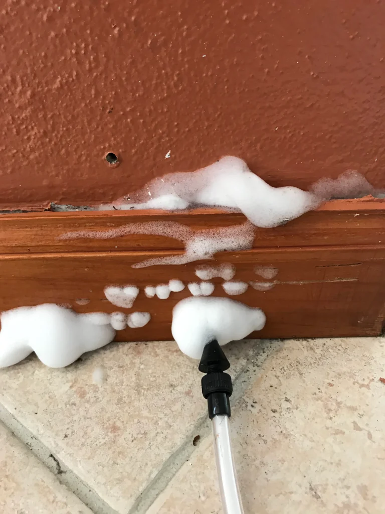 White foam is being injected into a wall via a tube and applicator near a brown baseboard for insulation.