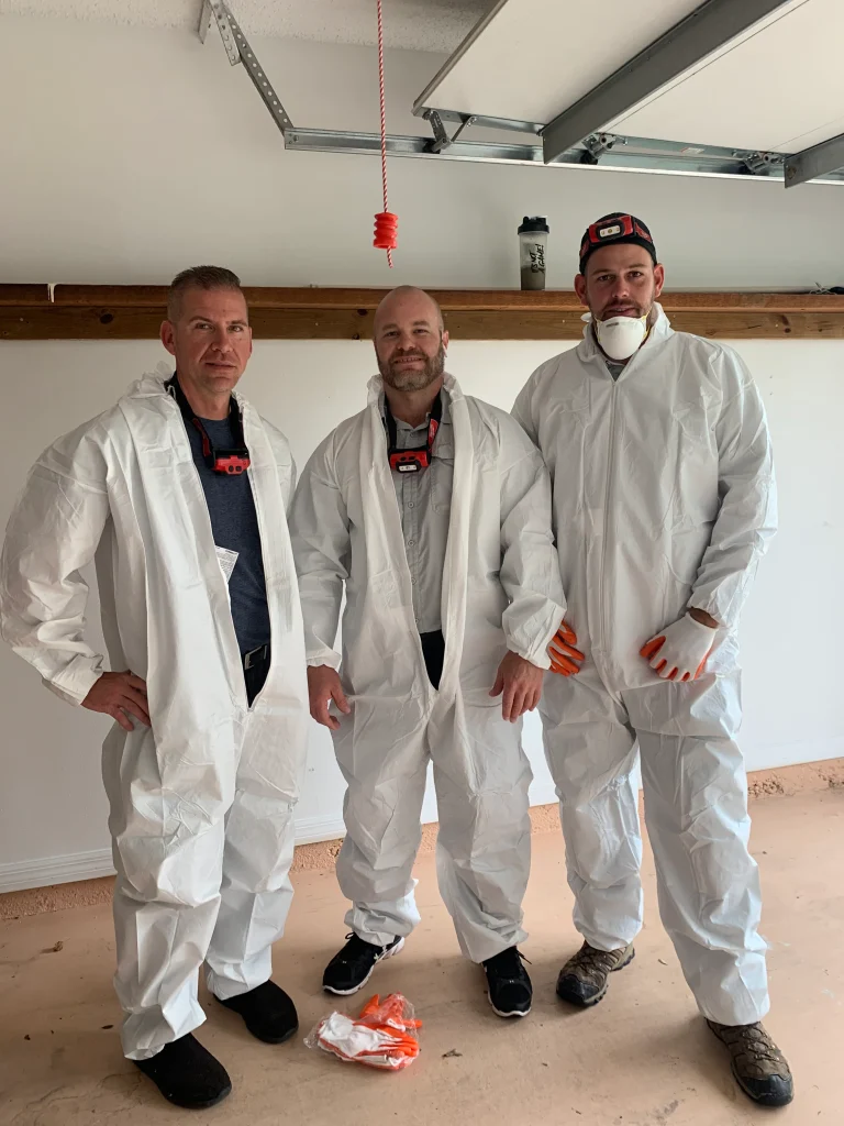 Three men in white protective coveralls and gloves stand together in a well-lit garage with light walls.