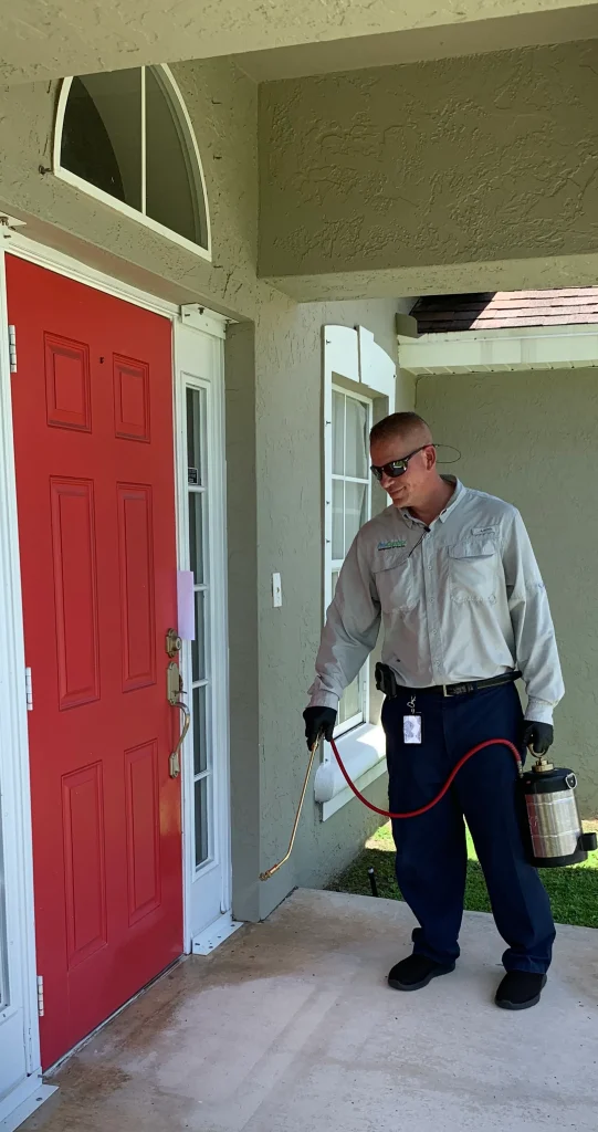 A pest control technician in gloves and sunglasses sprays pesticide outside a home’s red front door.